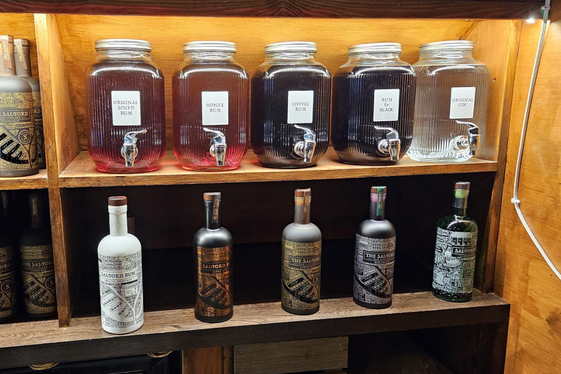 Salford Distillery bottle refill station at Arch 33 Salford, showing five dispensing jars of Original Spiced Rum, Honey Rum, Coffee Rum, Rum & Black, and Original Gin alongside the full bottled range